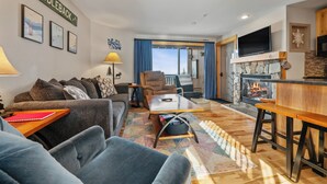 A living room with a sofa, armchairs, a coffee table, and a fireplace. There is a television mounted above the fireplace and a sign that reads "SADDLEBACK". - South Branch Condo 21-1 - ground level ski-in/out on Saddleback (Sandy River Plantation)