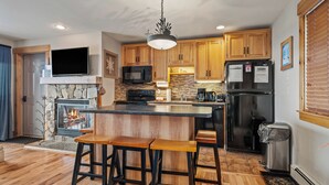 A modern kitchen with wooden cabinets, a built-in microwave, a stainless steel refrigerator, a stone fireplace, and a bar area with stools. - South Branch Condo 21-1 - ground level ski-in/out on Saddleback (Sandy River Plantation)