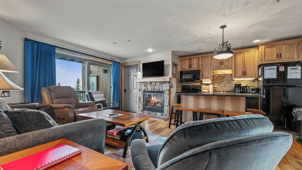 A modern living room with a fireplace, a television, and a kitchen area with wooden cabinets and a stone backsplash. - South Branch Condo 21-1 - ground level ski-in/out on Saddleback (Sandy River Plantation)