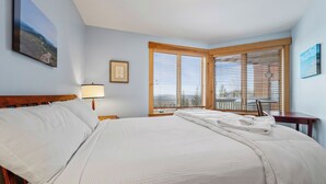 A neatly made bed with white linens, a bedside lamp, and a view of the outdoors through large windows with blinds. - South Branch Condo 21-1 - ground level ski-in/out on Saddleback (Sandy River Plantation)