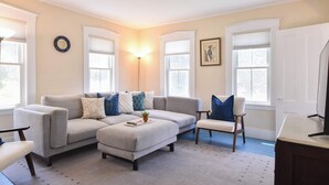 A living room with a grey sectional sofa, a small table with a plant, and a chair. There are two windows with white blinds, a wall clock, and a framed picture. - Private Downtown Wellfleet Location! (Wellfleet)