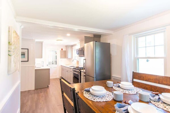 A modern kitchen with white cabinetry, a stainless steel refrigerator, and a dining area with a wooden table set for four. - 3 Min Walk to Beach! Large Yard! (West Yarmouth)