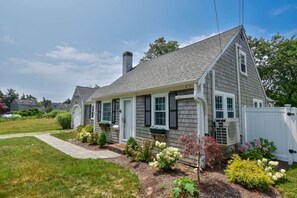 A two-story house with a grey shingle roof, a white front door, and a small porch with potted plants.