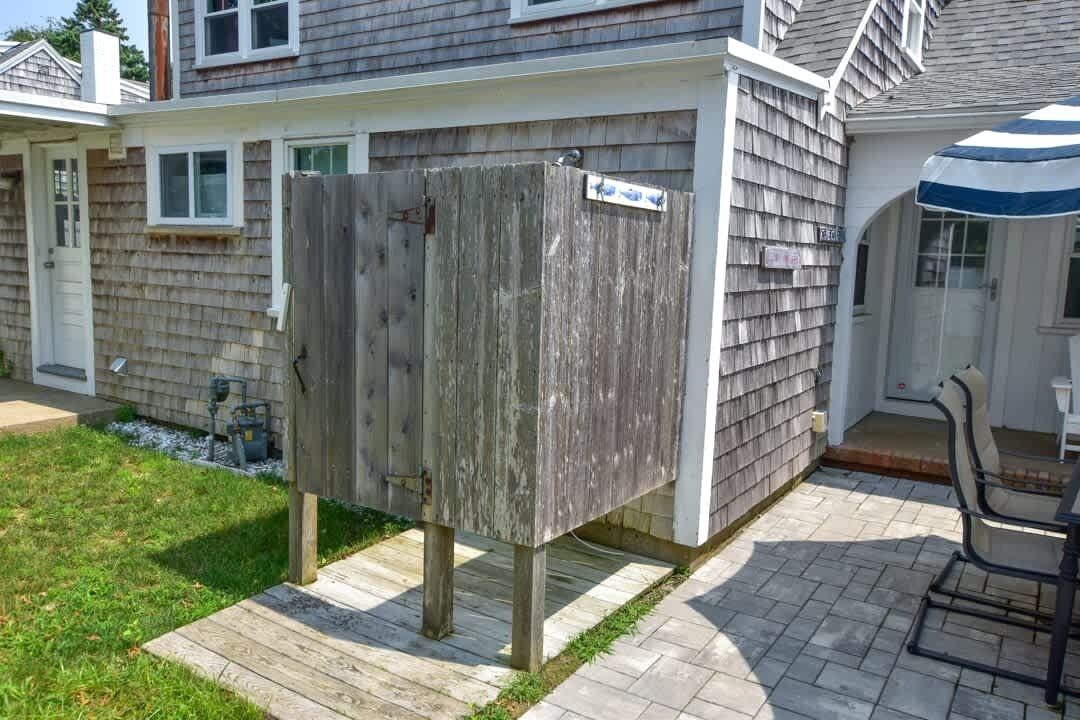 A wooden outdoor shower enclosure in a residential backyard with a patio and a chair.
