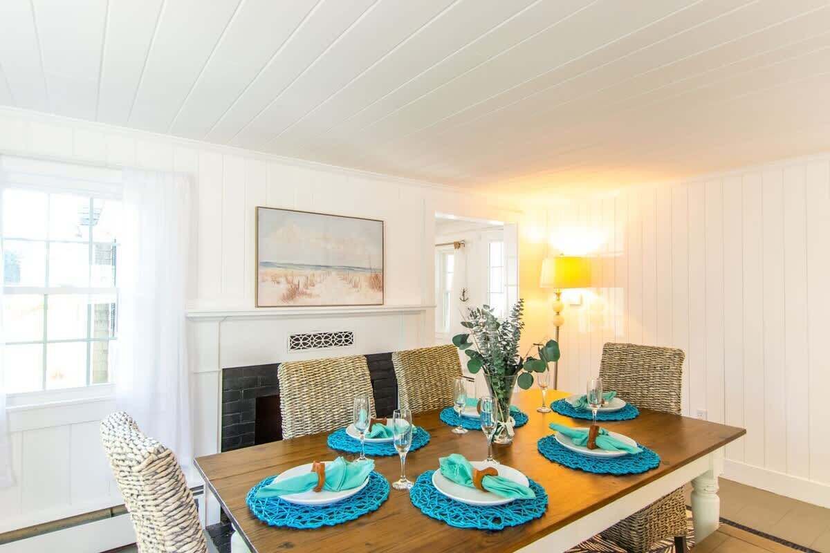 Un coin repas avec une table en bois dressée pour quatre personnes, entourée de chaises en osier, d’une cheminée et d’un tableau accroché au mur.