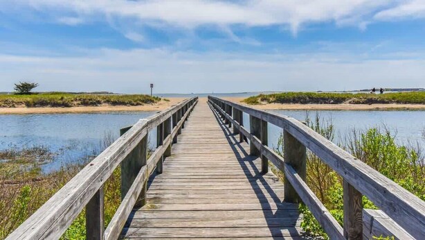 Une promenade en bois menant à un plan d’eau, entourée de zones herbeuses et sous un ciel dégagé.