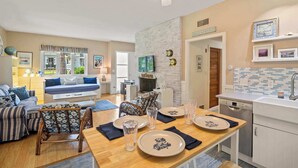 A well-lit living room with a sofa, a dining table set for four, and a kitchen area with a sink and cabinets. - Updated Beach House Deck Outdoor Shower! (Chatham)