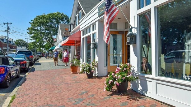A storefront with a sign reading "Saint New," a brick sidewalk, parked cars, and a flag. - Updated Beach House Deck Outdoor Shower! (Chatham)
