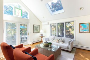 A spacious living room with a glass coffee table, a white sofa, and orange chairs. There are large windows with views of trees and a ceiling skylight. - Steps to Skaket Beach! (Orleans)