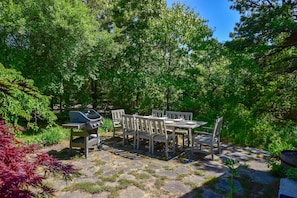 An outdoor dining area with a grill, stone pathway, and surrounding greenery. - Steps to Skaket Beach! (Orleans)