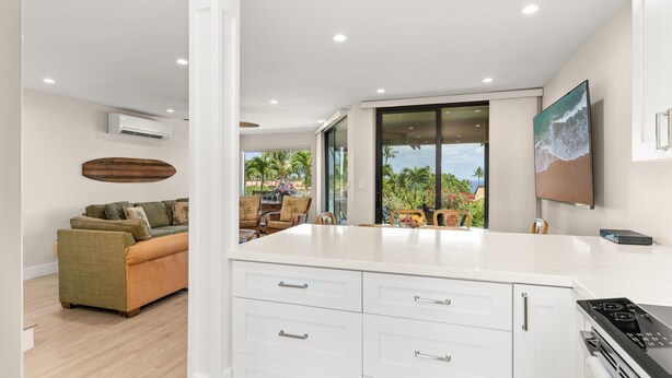 A modern kitchen with white cabinetry, a built-in stove, and a large window overlooking a patio with outdoor furniture. - Beautifully remodeled ocean view condo in Wailea WE27B (Kihei)