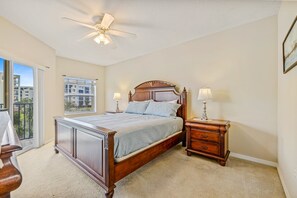 Een slaapkamer met een groot bed, een nachtkastje, een lamp, een plafondventilator en een raam met uitzicht op gebouwen.