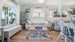 A room with a sofa, a small table, a ceiling fan, a window with curtains, and a view of palm trees.