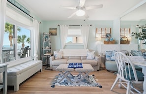 A room with a sofa, a small table, a ceiling fan, a window with curtains, and a view of palm trees.