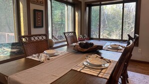 Un coin repas avec une table en bois dressée pour quatre personnes, de grandes fenêtres et une vue sur les arbres.