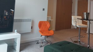 Una habitación de hotel moderna con un televisor de pantalla plana, una silla de oficina naranja, una silla blanca, un reposapiés verde y una puerta de madera.