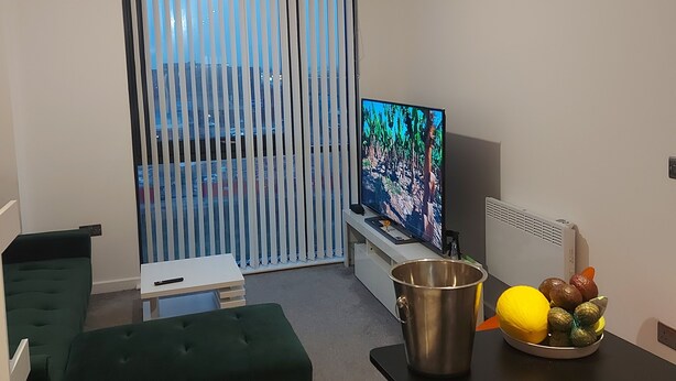 Una sala moderna con un televisor de pantalla plana, un otomano verde, una mesa de centro con frutas y un ventanal corredizo que da a un balcón.