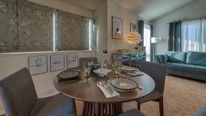 Un coin repas avec une table ronde dressée pour quatre personnes, entourée de chaises. Des œuvres d’art encadrées ornent le mur, une fenêtre est agrémentée de stores à motifs floraux, et un canapé se trouve au fond de la pièce.