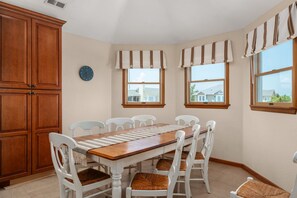 Un coin repas avec une table en bois et des chaises blanches, de grandes armoires en bois et des fenêtres ornées de rideaux à rayures.