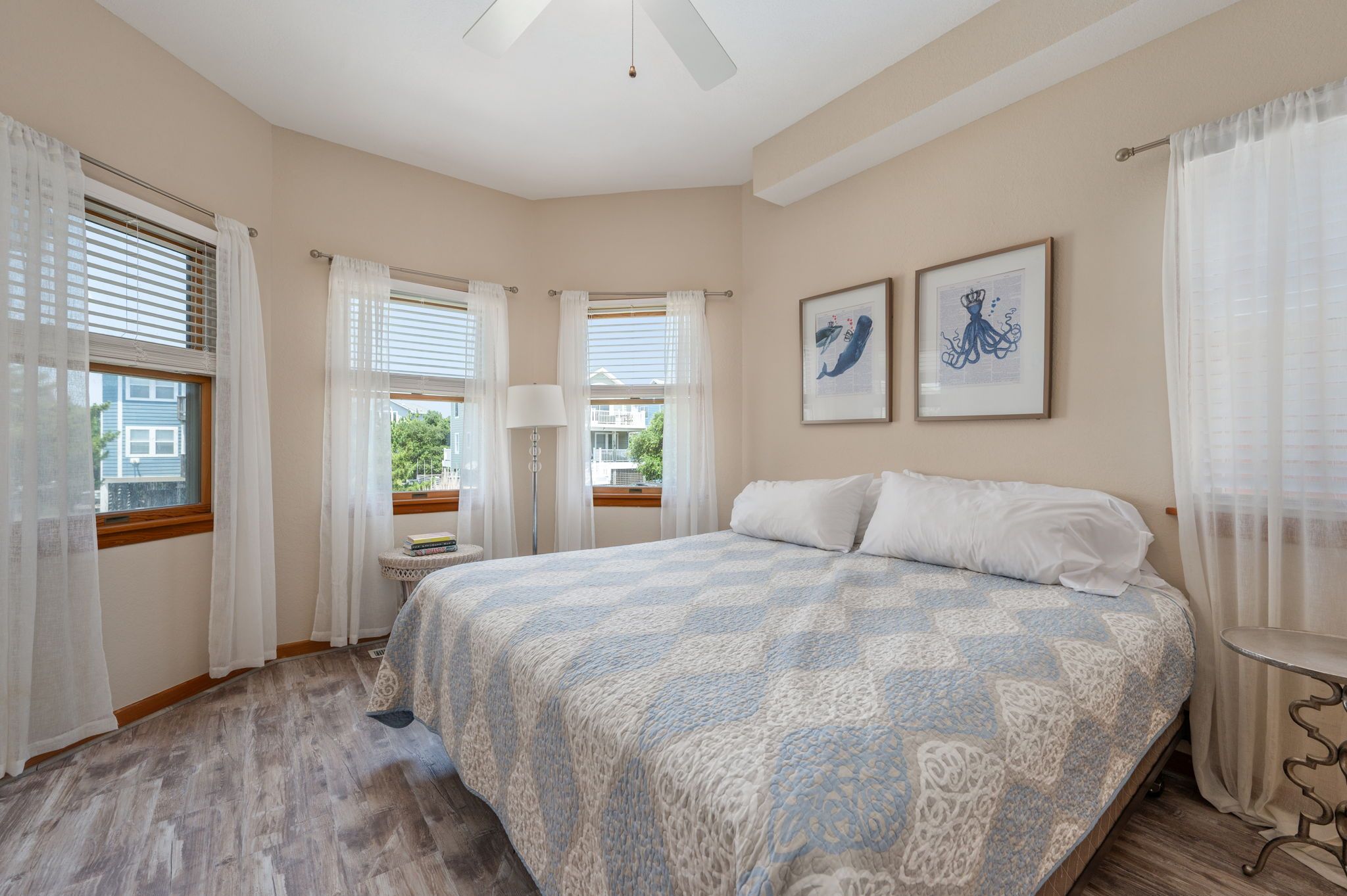 Un dormitorio con una cama grande, dos ventanas con cortinas, una mesita de noche con lámpara y dos cuadros enmarcados en la pared.