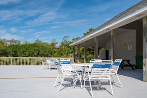 A covered outdoor seating area with white metal chairs and tables, a wooden deck, and a view of trees and sky. - The Purple Dandelion-Oceanside,dog-friendly 🐶easy beach access,community pool (Corolla)