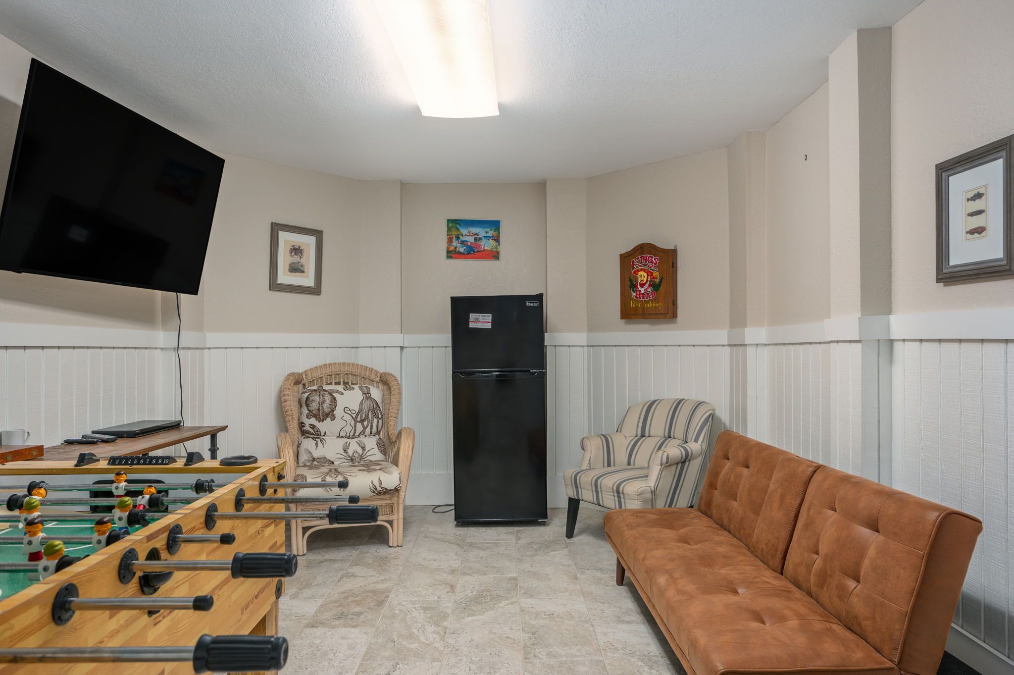 A living room with a foosball table, two armchairs, a sofa, a flat-screen TV, and a refrigerator.