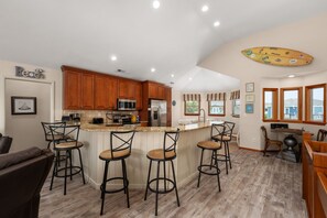 A kitchen with wooden cabinets, a central island with bar stools, and a large window with a view. - The Purple Dandelion-Oceanside,dog-friendly 🐶easy beach access,community pool (Corolla)