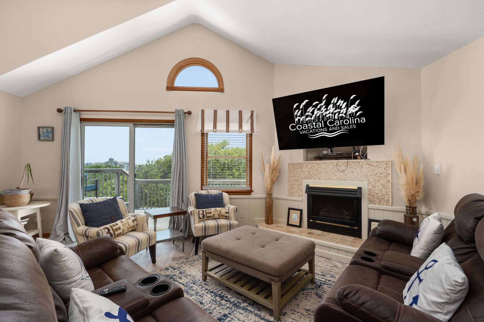A spacious living room with a fireplace, comfortable seating, and a large TV screen displaying 'Coastal Carolina Vacations and Sales'.