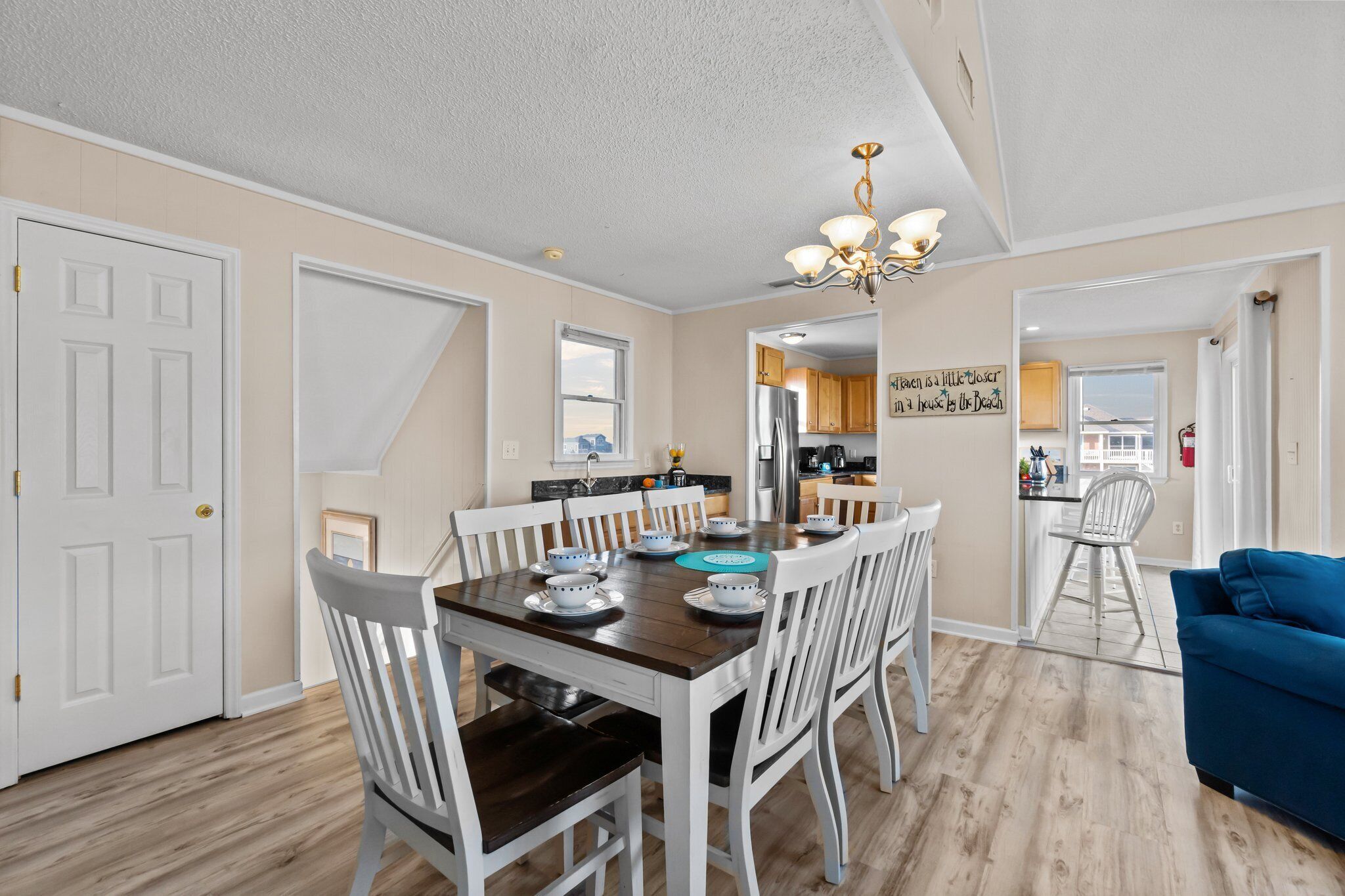 A dining area with a wooden table set for six, white chairs, a chandelier, and a view of the kitchen in the background.