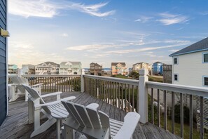 Une terrasse en bois avec des chaises Adirondack blanches, donnant sur une rangée de maisons de plage, sous un ciel dégagé.