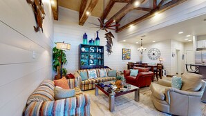 Un salón espacioso con un sofá grande, una mesa de centro y un sillón rojo. Se observa una estantería con objetos decorativos y un comedor al fondo.