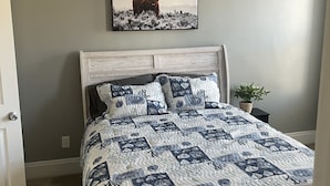 Un dormitorio con una cama, un ventilador de techo, un cuadro en la pared y una pequeña planta en una mesita de noche.