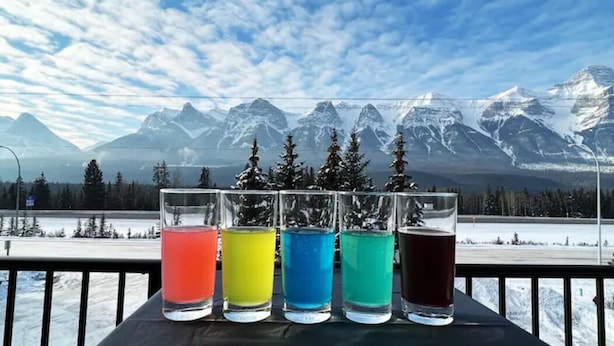 A table with five glasses of colored liquids, set against a backdrop of snow-covered mountains and a clear sky. - NewReno Spectacula MountainView KingBed Jacuzzi Chalet (Harvie Heights)