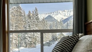 A bed with patterned pillows, a window with a view of snow-covered trees and mountains, and a glimpse of a street with parked cars. - NewReno Spectacula MountainView KingBed Jacuzzi Chalet (Harvie Heights)