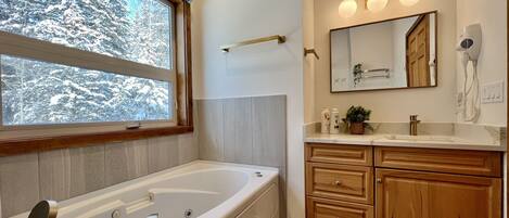 Un baño moderno con una bañera grande, muebles de madera, un tocador con espejo y una ventana con vistas a árboles cubiertos de nieve.
