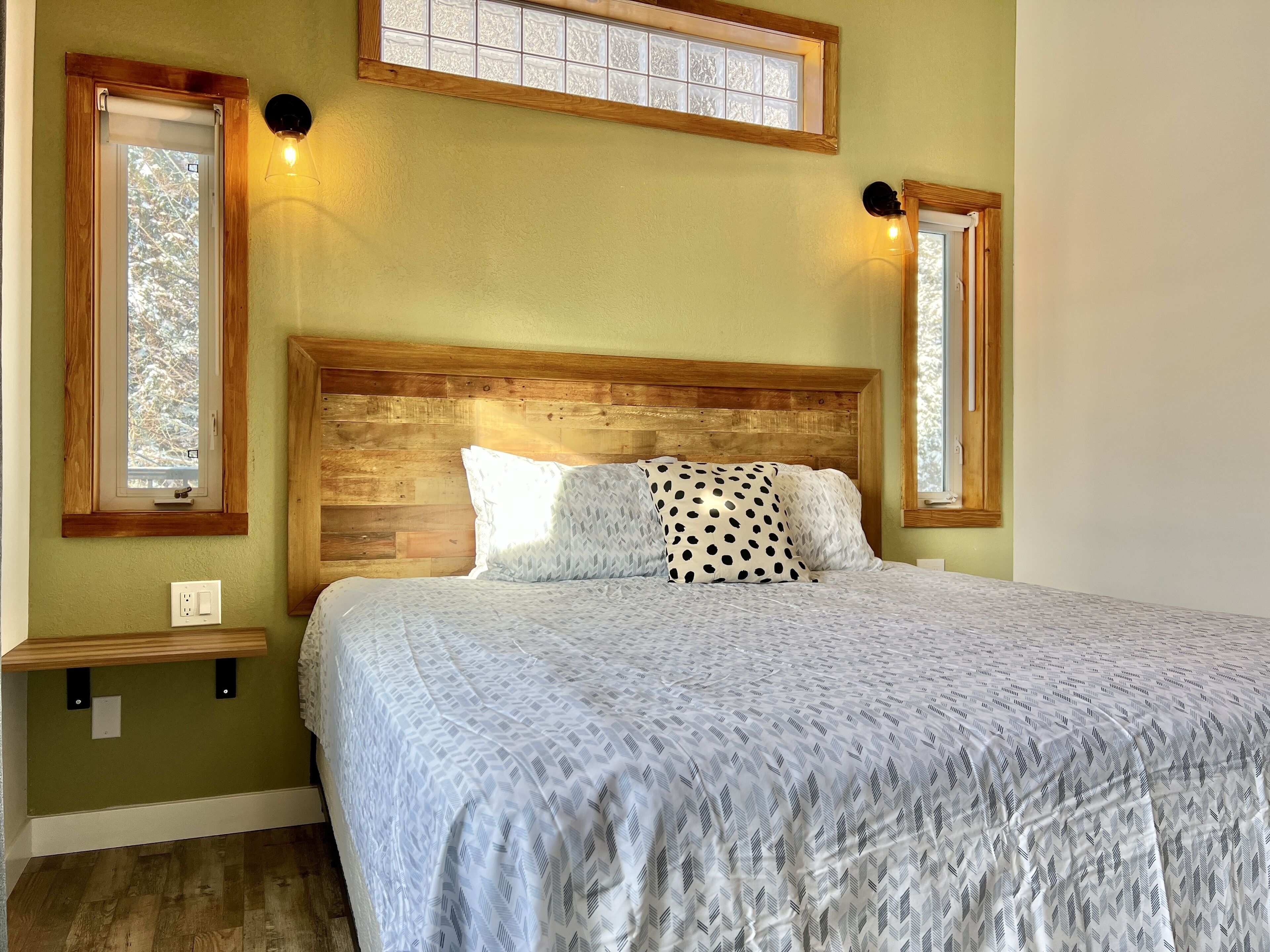A bedroom with a wooden headboard, a bed with patterned bedding, two wall-mounted lights, and a window with a view of trees.