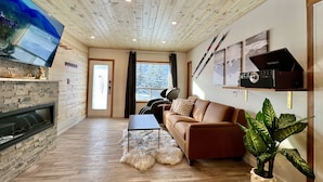 A cozy living room with a stone fireplace, a brown leather sofa, a flat-screen TV, and ski equipment on the wall. - NewReno Spectacula MountainView KingBed Jacuzzi Chalet (Harvie Heights)