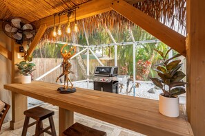 Um balcão de madeira com grelha e plantas em vasos, com vista para um jardim com cerca de madeira e plantas tropicais.