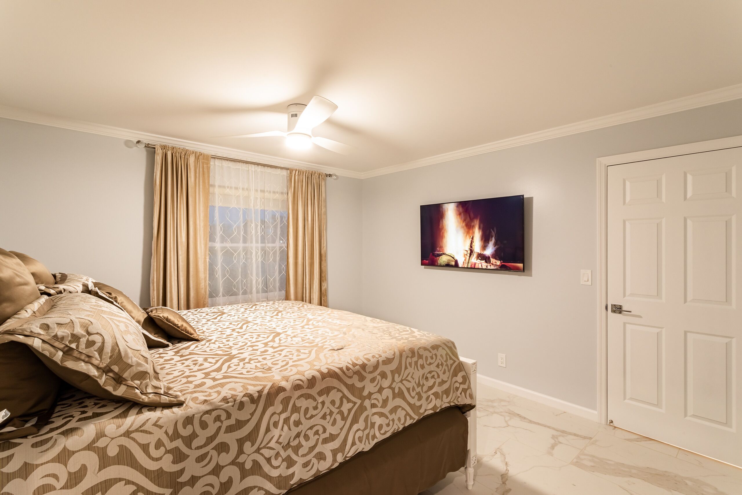 A bedroom with a bed, a ceiling fan, a window with curtains, a door, and a wall-mounted TV.