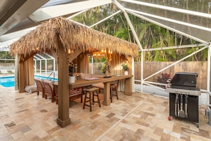 An outdoor patio with a thatched-roof gazebo, a wooden table with chairs, a grill, and a pool in the background.
