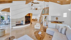 Ein großzügiger Wohnbereich mit Kamin, weißen Sofas, einem Holztisch und einem Deckenventilator.