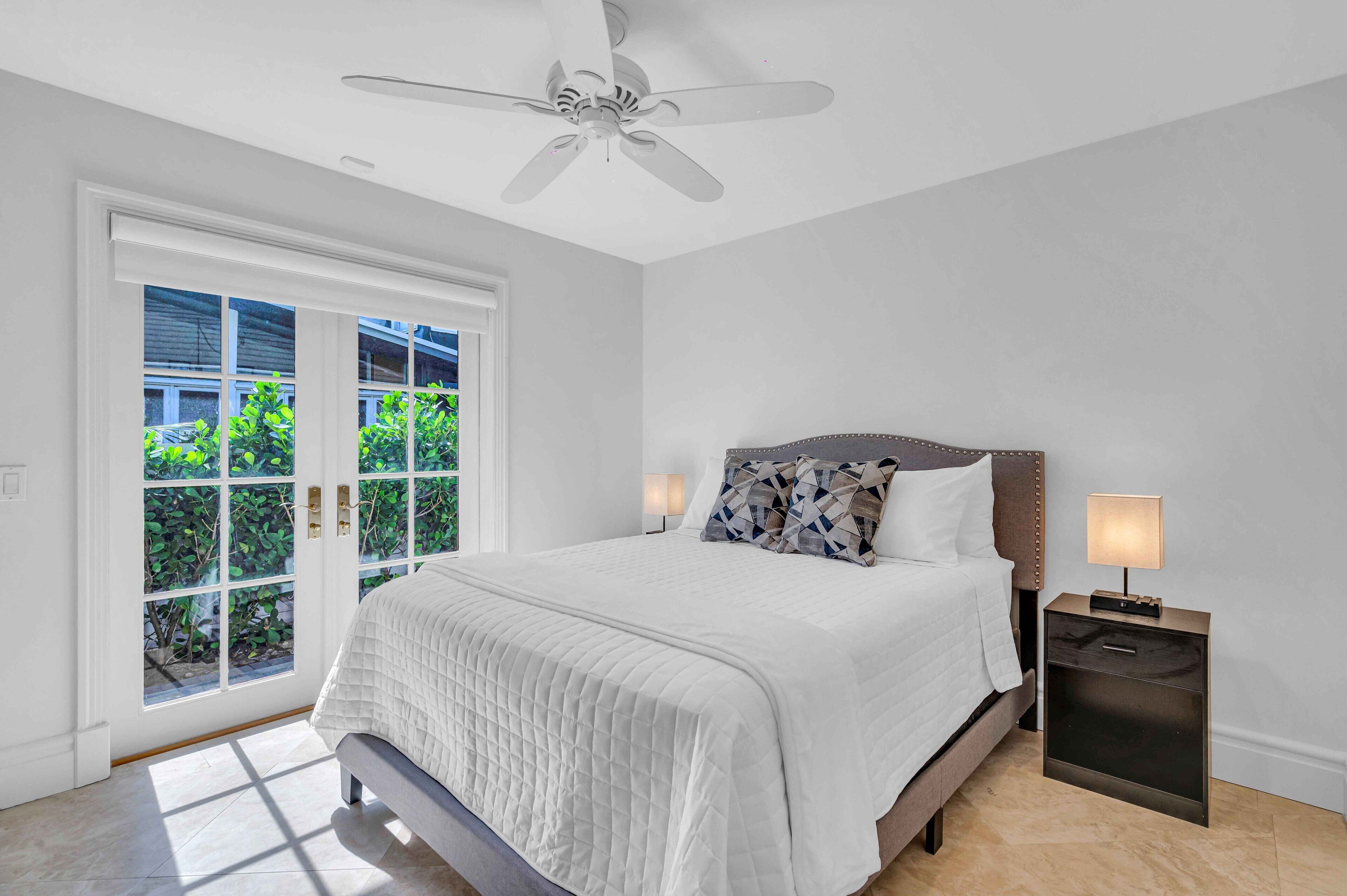 A bedroom with a bed, a nightstand, a ceiling fan, and a view of greenery through a sliding door.