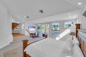 Quarto com uma cama grande, uma escrivaninha e uma cadeira, escada que leva a uma varanda, e janelas amplas com vista para palmeiras e uma casa.