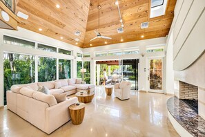 A spacious living room with a large sectional sofa, a fireplace, and a view of the outdoors through large windows. - Architectural Masterpiece | Venice of America | Riverside View & Access | Nurmi Key | RESlDENCES (Fort Lauderdale)