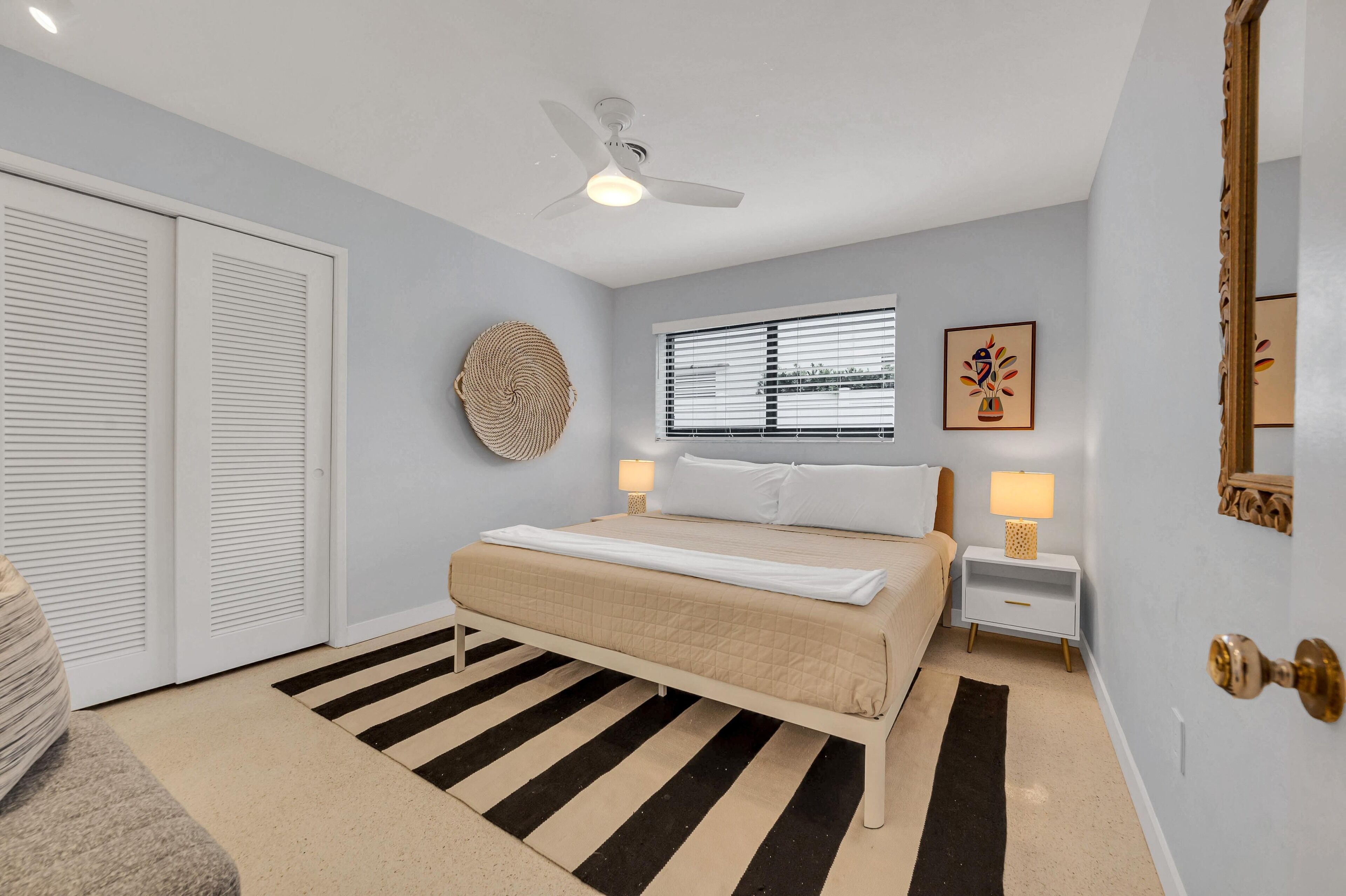 A bedroom with a bed, bedside lamps, a ceiling fan, a window with blinds, a wall clock, and a door with a brass handle.