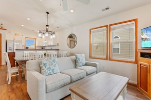 A living room with a sofa, coffee table, and a television. There is a kitchen area with cabinets and a refrigerator.