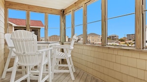 A covered porch with wooden chairs and a table, large windows, and a view of surrounding buildings. - 5180 Perfect Pastime * 5 Min Walk to Beach (Kill Devil Hills)