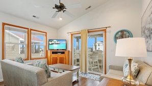 A spacious living room with a television, a ceiling fan, and a view of the ocean through large windows. - 5180 Perfect Pastime * 5 Min Walk to Beach (Kill Devil Hills)