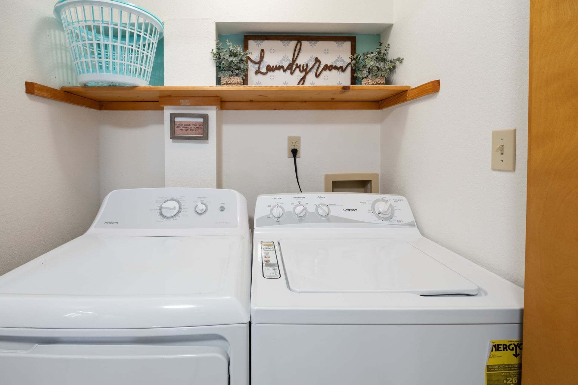 A laundry room with two white front-loading washing machines, a blue laundry basket, a wooden shelf with a 'Laundry Room' sign, and a small framed sign on the wall.