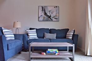 Un salón con un sofá azul, una mesa de centro con libros, una lámpara y un cuadro en la pared.